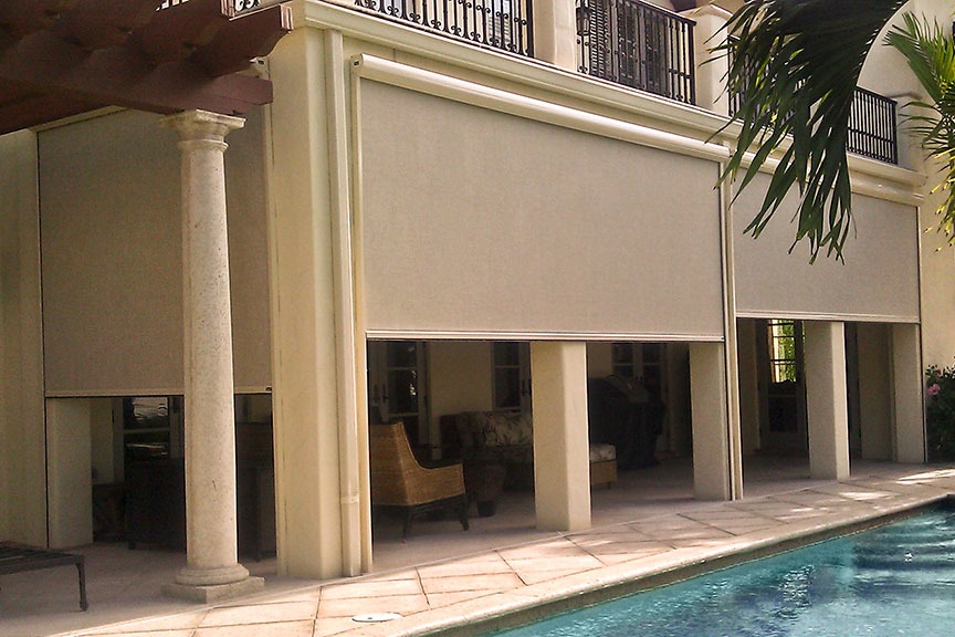 A set of beige Sunesta Sentry retractable screens, providing poolside shade for an outdoor seating area.